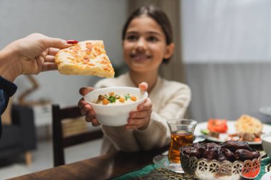 Muslim woman holding pita bread near blurred daughter with sauce during iftar at home 