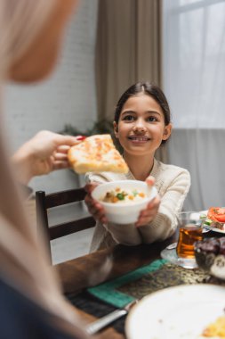 Positive muslim girl holding sauce near blurred mom with pita bread at home 