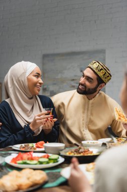 Smiling man holding pita bread and looking at wife in hijab during iftar at home 