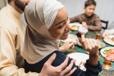 Muslim man hugging wife in hijab holding date fruit at home 