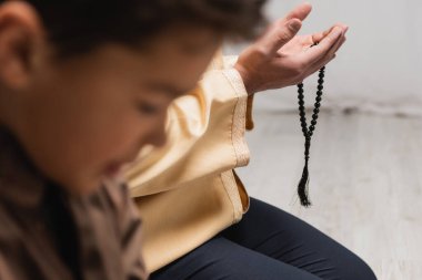 Muslim man with prayer beads praying near blurred son at home 
