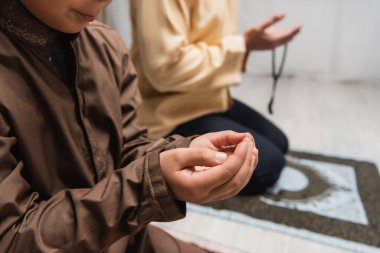 Cropped view of muslim boy praying during salah near blurred dad at home 