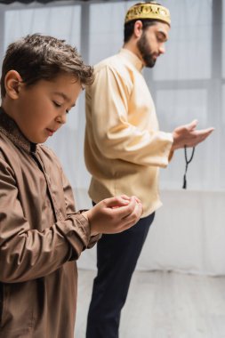 Muslim boy praying near blurred father during ramadan at home 