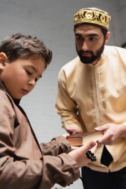 Blurred muslim father giving book to son with prayer beads at home 