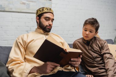 Muslim man reading book near son on couch at home 