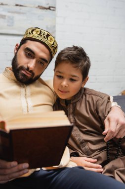 Müslüman bir adam oğlunu kucaklıyor ve evde kitap okurken dua boncukları tutuyor. 