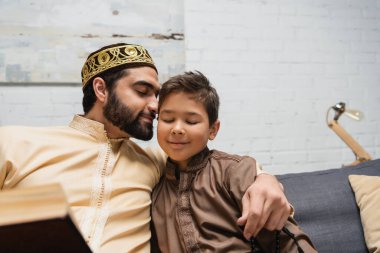 Gülümseyen Müslüman adam, dua boncuklarıyla evdeki kanepenin yanında oğluna sarılıyor. 