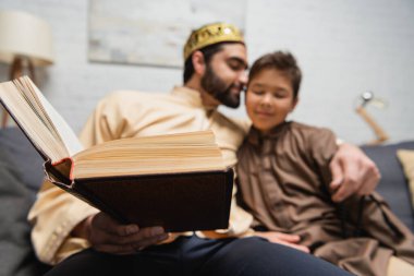 Book in hand of muslim man hugging son on couch at home  clipart