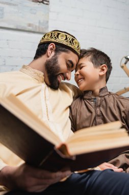 Cheerful middle eastern man and son reading blurred book at home 