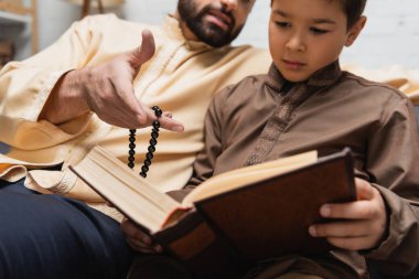 Muslim man with prayer beads pointing at book near son at home 