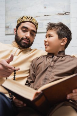 Arabian man holding prayer beads while talking to son with blurred book at home 