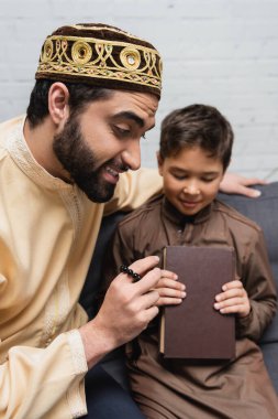 Dua boncuklarıyla gülümseyen müslüman, oğlunun yanında elinde kitapla oturuyor. 