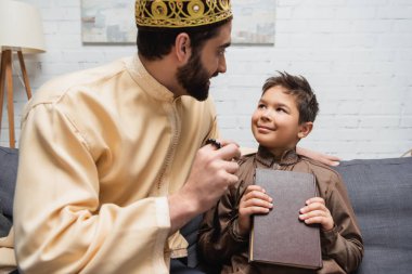 Muslim father talking to son holding koran on couch at home 