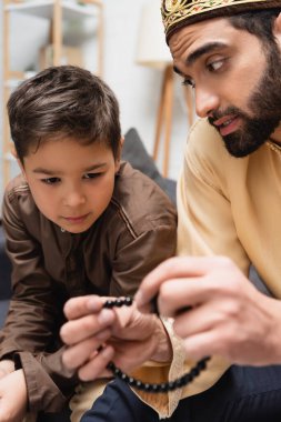 Middle eastern man holding blurred prayer beads and talking to son at home 