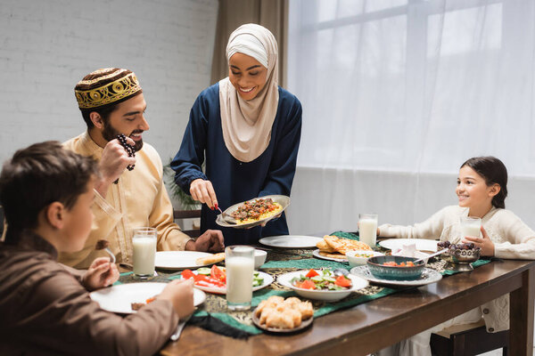 African american woman in hijab serving food on plate near family and ramadan dinner 
