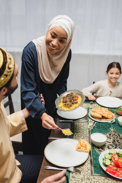 African american woman in hijab serving pilaf on plate near husband during ramadan at home 