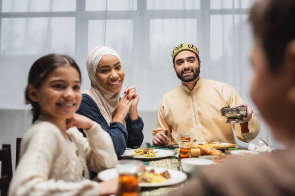Positive middle eastern family looking at blurred boy during ramadan dinner 