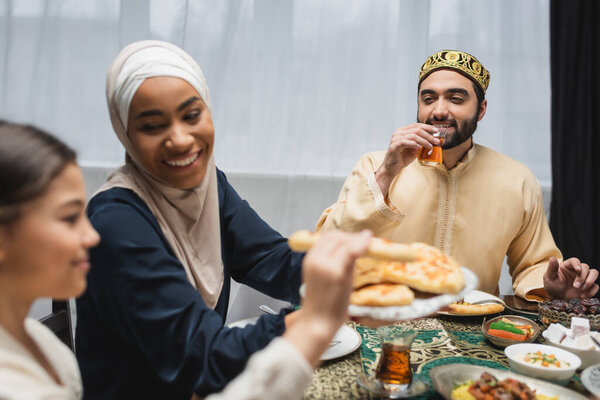 Smiling muslim man holding turkish tea glass near family and ramadan dinner 