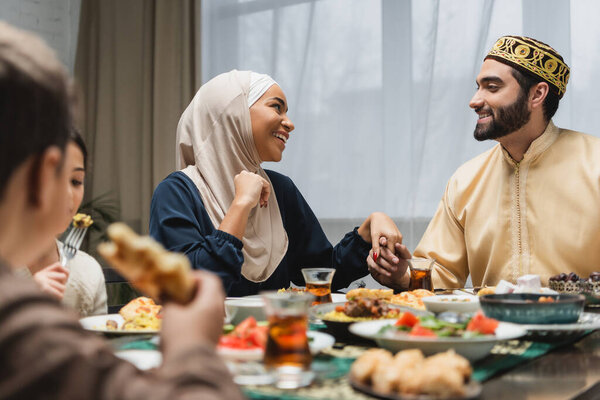 Smiling muslim family holding hands near kids and ramadan dinner 