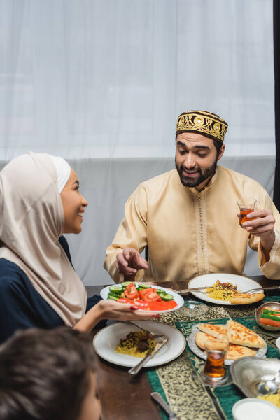 Smiling muslim man holding tea near wife and food during iftar at home 