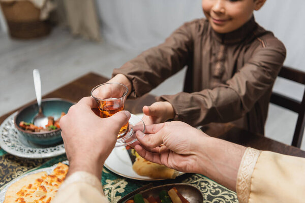 Cropped view of muslim man giving turkish tea glass to son during iftar at home 
