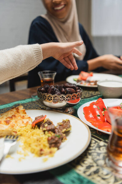 Обрезанный вид мусульманского ребенка, указывающего пальцем на размытую мать и еду дома 