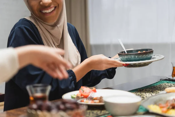 Cropped view of smiling muslim woman holding food near daughter at home