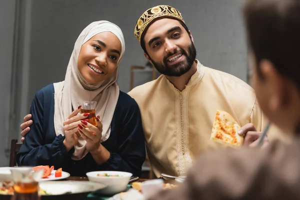 Smiling muslim parents holding food and tea near blurred child at home