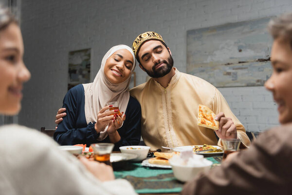 Arabian man hugging wife in hijab near blurred kids during iftar at home 