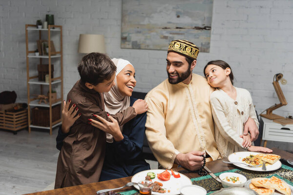 Cheerful muslim parents hugging kids near dinner during ramadan at home 