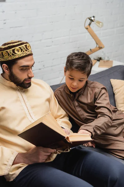 Musulmán Chico Leyendo Libro Con Padre Sofá Casa — Foto de Stock