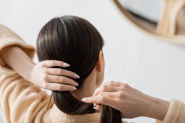 back view of young brunette woman adjusting shiny hair near mirror in bathroom