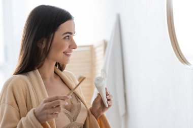 side view of happy young woman holding tube with toothpaste and toothbrush while looking at mirror 