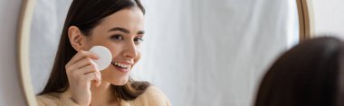 happy young woman cleansing face with cotton pad and looking at mirror in bathroom, banner 