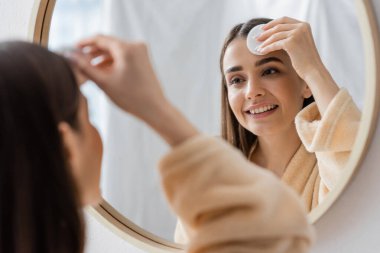 Gülümseyen genç bir kadının banyo aynasında pamuk yastıkla yüzünü temizlemesinin yansıması. 