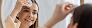 reflection of smiling young woman cleansing face with cotton pad in bathroom mirror, banner 