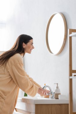 side view of young happy woman with brunette long hair looking at mirror in bathroom 