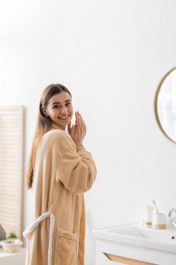 positive woman in bathrobe looking at camera near mirror in bathroom 