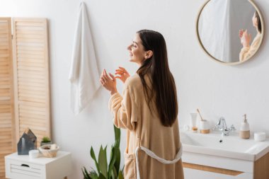positive woman in bathrobe laughing near mirror in modern bathroom 