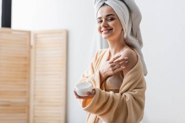 pleased young woman with towel on head holding container while applying body butter 