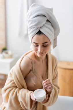 young woman in bathrobe with towel on head holding container with body butter 