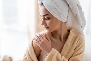 young woman in bathrobe with towel on head applying body cream in bathroom 