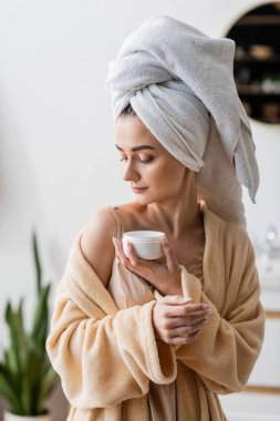 Young woman in towel on head and bathrobe holding cosmetic cream 
