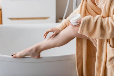 Cropped view of young woman applying cosmetic cream on leg near bathtub 