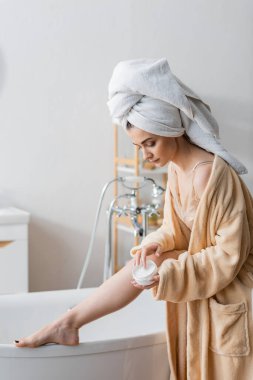 Young woman in towel and bathrobe holding body cream near bathtub at home 