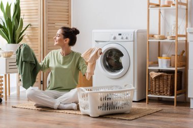 Side view of cheerful brunette woman holding clothes near basket in laundry room 