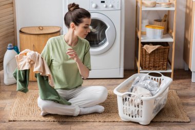 Side view of young woman holding colorful clothes near basket in laundry room 
