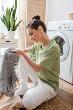 Side view of young woman holding t-shirt near clothes and washing machine at home 