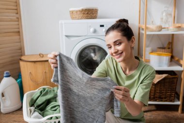 Positive brunette woman holding blurred t-shirt in laundry room 