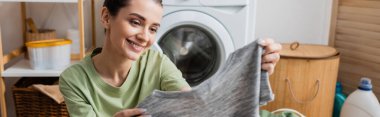 Smiling woman holding t-shirt in laundry room, banner 